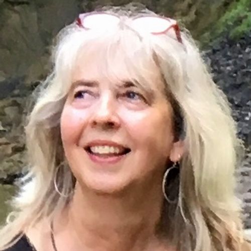A woman with long gray-blonde hair smiles as she looks slightly upward. She is wearing hoop earrings and glasses resting on her head. The background is softly blurred, showing an outdoor setting with greenery.